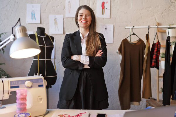 Photo of long-haired seamstress woman with arms crossed at waist standing next to mannequin.