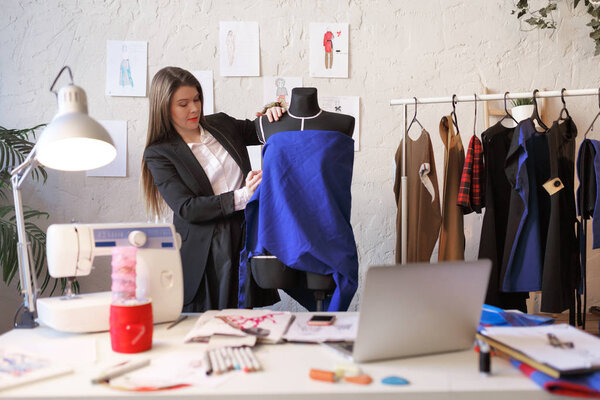 Photo of long-haired seamstress woman standing next to mannequin with blue cloth.