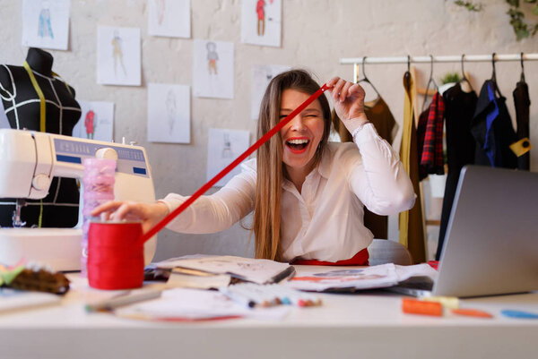 Photo of happy female dressmaker unwinding red ribbon sitting at table in atelier