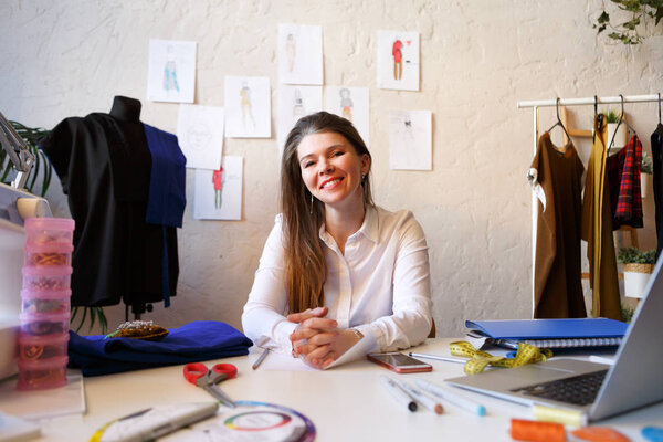 Photo of female dressmaker sitting at table with sewing machine and laptop