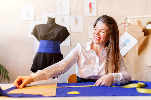 Photo of happy dressmaker female sitting at table with cloth, piece, tape measure.
