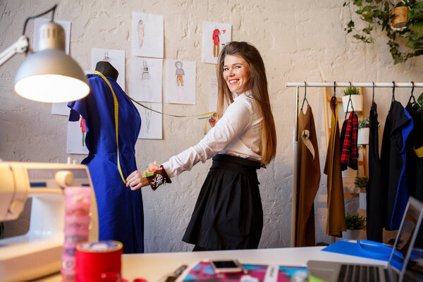 Image of female dressmaker with phone in hands standing next to mannequin with blue cloth.
