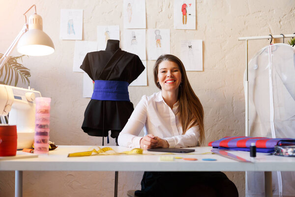 Photo of happy dressmaker female sitting at table with cloth, tape measure.