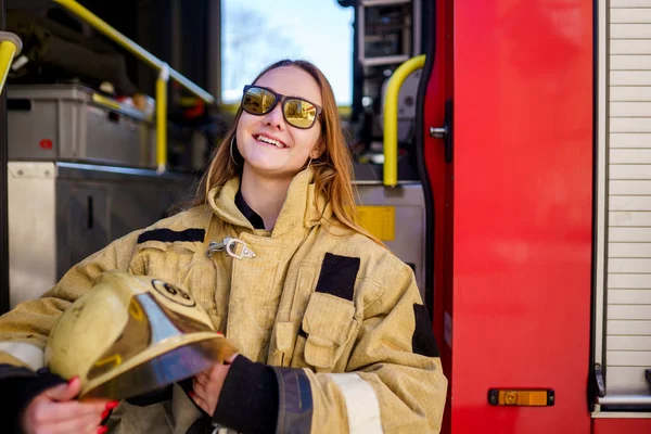 Foto de mujer de fuego sonriente en gafas con casco en las manos sentado en camión de bomberos — Foto de Stock