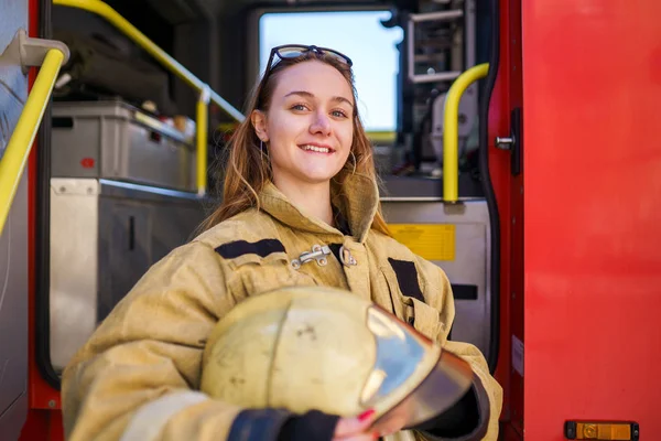 Imagen de mujer bombero sonriente con gafas en la cabeza con casco en las manos de pie junto al camión de bomberos — Foto de Stock