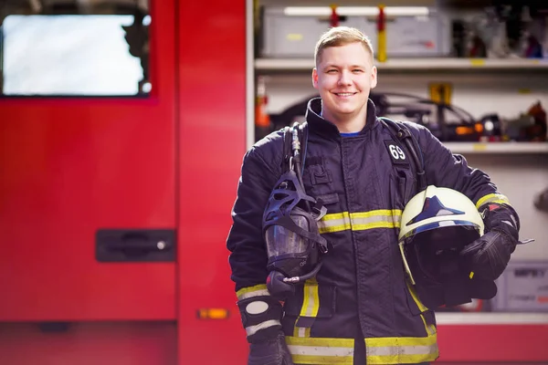 Foto de feliz bombero masculino con casco en las manos contra el fondo del camión de bomberos — Foto de Stock