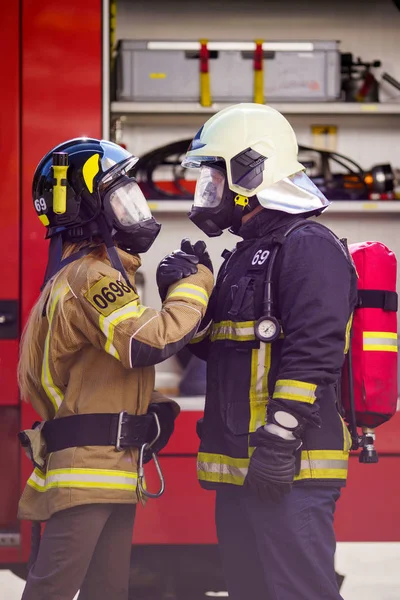 Imagem de bombeiros mulheres e homens no capacete e máscara olhando uns para os outros e fazendo aperto de mão perto caminhão de bombeiros — Fotografia de Stock