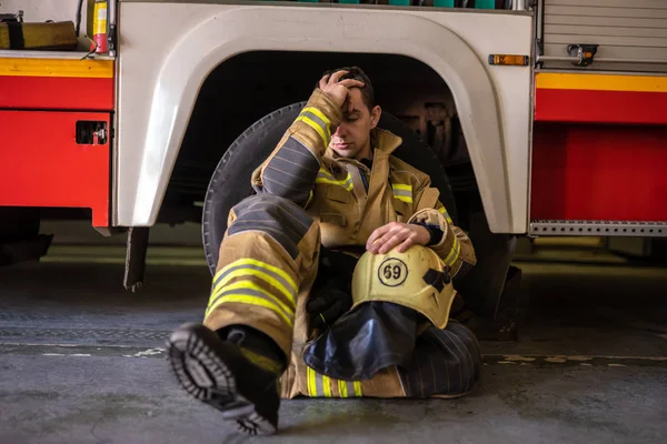 Foto eines jungen müden Feuerwehrmannes, der auf dem Boden neben einem Feuerwehrauto sitzt — Stockfoto