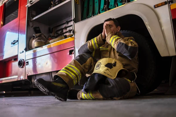 Foto de bombero cansado sentado en el suelo cerca de camión de bomberos rojo —  Fotos de Stock