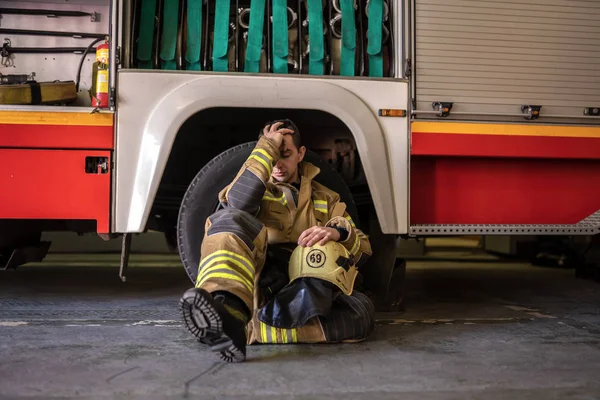 Зображення втомленого пожежника, що сидить на підлозі біля пожежної машини — стокове фото