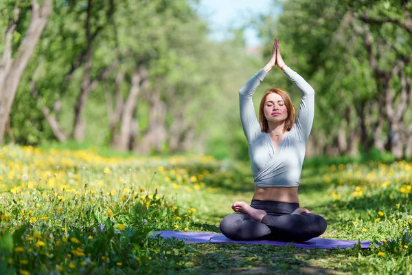 Foto de una mujer haciendo yoga con los brazos levantados sentada en posición de loto sobre una alfombra azul en el bosque —  Fotos de Stock