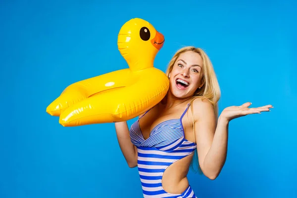 Photo of happy blonde in striped swimsuit with lifebuoy duckling in hand on empty blue background — Stock Photo, Image