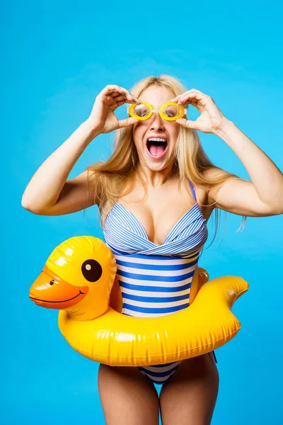 Image of screaming blonde in striped swimsuit, swimming goggles with lifebuoy duckling at waist on empty blue background — Stock Photo, Image