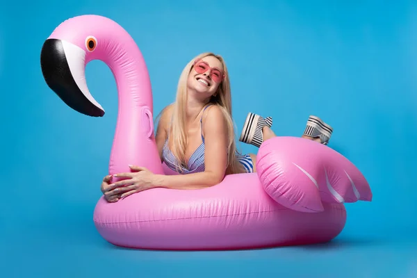 Photo of blonde girl in bathing suit and in pink glasses lying on pink inflatable flamingo on empty blue background — Stock Photo, Image