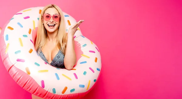 Boş pembe arka plan üzerinde yüzmek için şişme donut ile mayo güneş gözlüğü kız Fotoğrafı. Elle yana gösterir — Stok fotoğraf