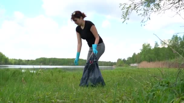 Woman picks up trash puts cups into bag on river meadow — ストック動画