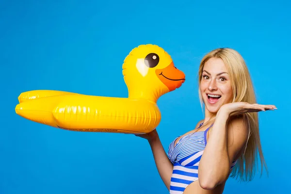 Photo of young blonde in striped swimsuit with lifebuoy duckling in hand on empty blue background — Stock Photo, Image