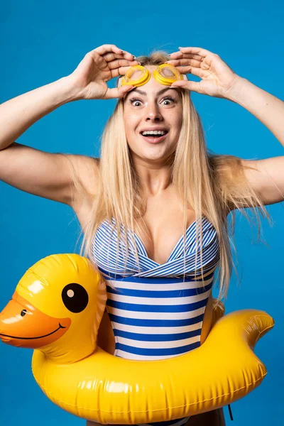 Photo of blonde in striped swimsuit, swimming goggles with lifebuoy duckling at waist on empty blue background — Stock Photo, Image