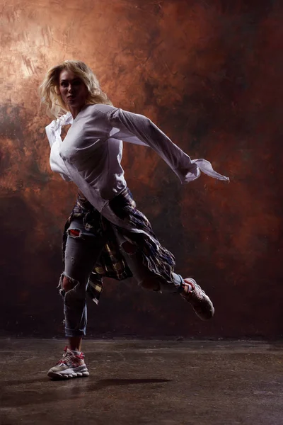Foto de jovem dançarina loira em jeans rasgados e tênis em fundo marrom escuro — Fotografia de Stock