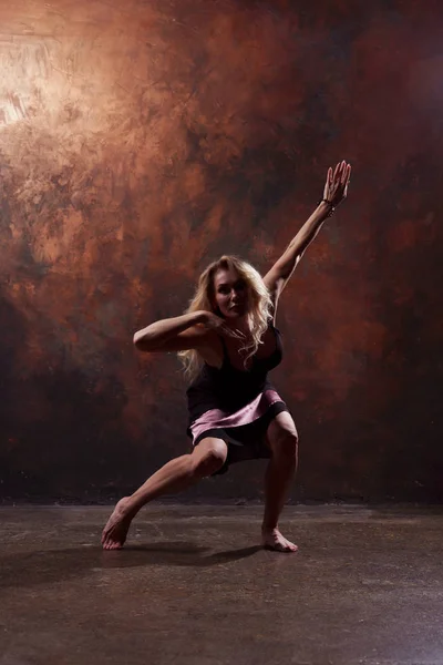 Foto de cuerpo entero de chica rubia bailando mirando en cámara en vestido negro corto sobre fondo marrón — Foto de Stock