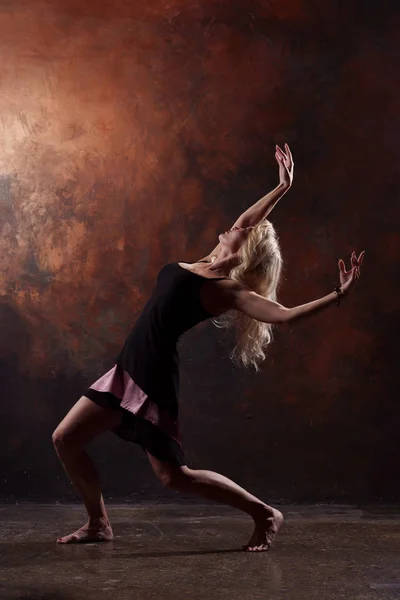 Foto de chica rubia bailando mirando hacia atrás en vestido negro sobre fondo marrón —  Fotos de Stock
