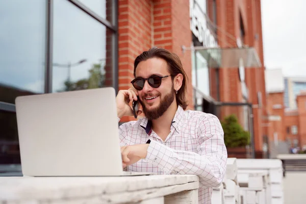 Foto di un uomo freelance sorridente che parla al telefono seduto a tavola vicino all'edificio in mattoni con laptop — Foto Stock