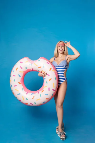 Foto de mujer rubia en traje de baño con rosquilla inflable sobre fondo azul vacío — Foto de Stock
