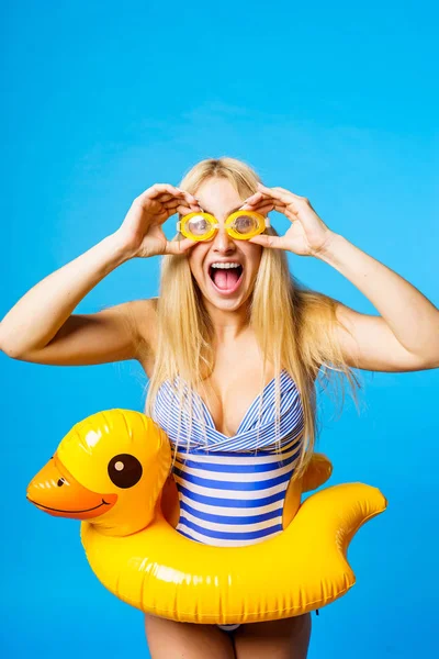 Photo of screaming blonde in striped swimsuit, swimming goggles with lifebuoy duckling at waist on empty blue background — Stock Photo, Image