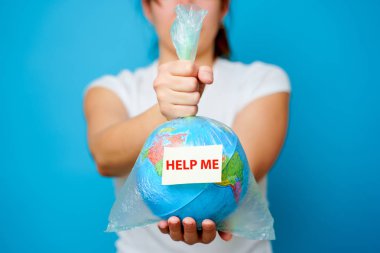 Woman holds in hands plastic bag with sticker text HELP ME and planet earth at blue background. The concept of plastic pollution. clipart