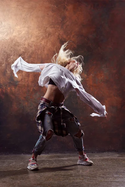 Zdjęcie smukłej blond tancerki z rozwojem włosów w rozdarty dżinsy i trampki na ciemnym tle brązowym — Zdjęcie stockowe