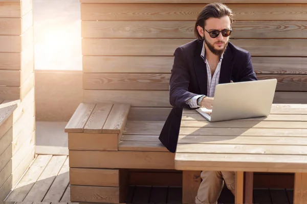 Foto di giovane uomo bruno con la barba in occhiali da sole seduto con computer portatile su una panchina di legno in primavera — Foto Stock