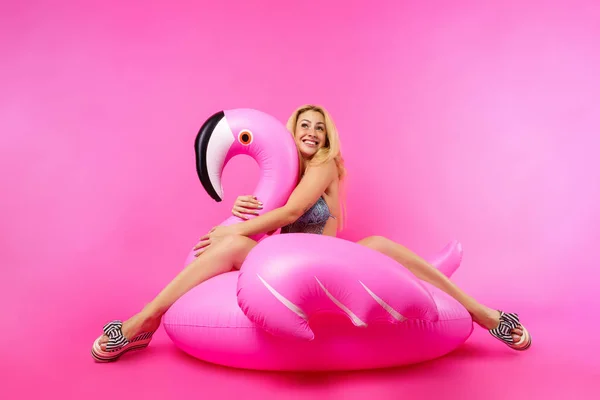Image of blonde girl in bathing suit and in pink glasses sitting on inflatable flamingo on empty pink background — Stock Photo, Image