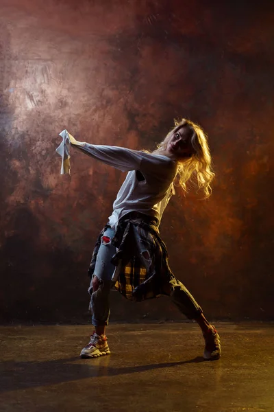 Foto de dança de cabelos longos menina loira olhando para a câmera em jeans rasgado no fundo marrom — Fotografia de Stock