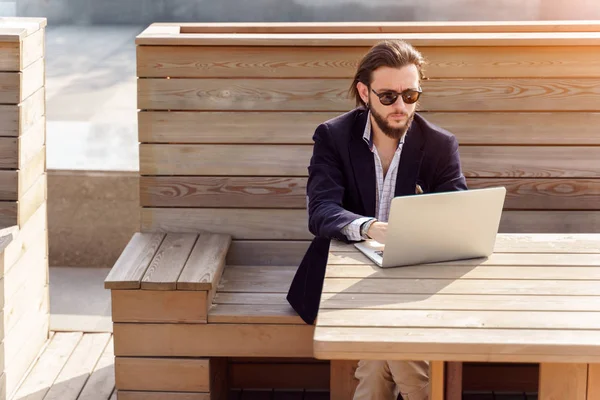 Foto di uomo bruno con la barba in occhiali da sole seduto con computer portatile su una panchina di legno in primavera — Foto Stock