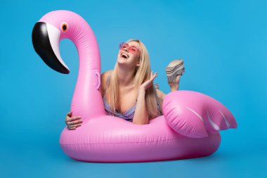 Boş mavi arka plan üzerinde pembe şişme flamingo üzerinde yatan mayo ve pembe gözlük sarışın resmi