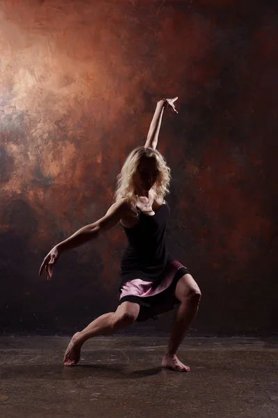 Foto av ung dansande blondin med upphöjda armar i svart klänning på brun bakgrund — Stockfoto