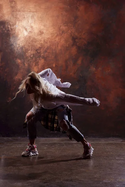 Foto de dançarina loira magra em jeans rasgados e tênis em fundo marrom escuro — Fotografia de Stock