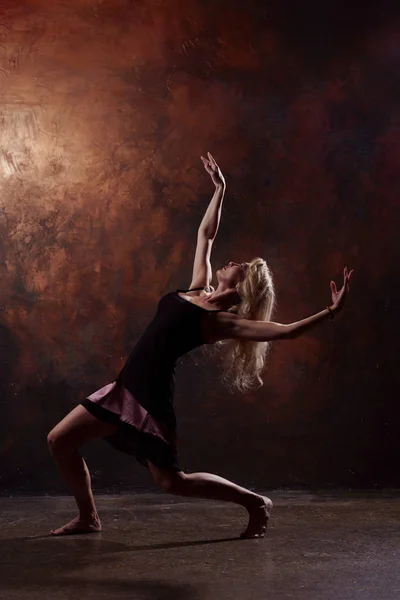 Foto de larga duración de la joven bailarina rubia mirando hacia arriba en vestido negro corto sobre fondo marrón —  Fotos de Stock