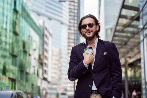 Fotó boldog Brunet férfi szakállas és fülbevaló fül üveg a kezében a városban a háttérben a sokemeletes épületek, — Stock Fotó