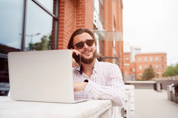 Foto de homem feliz freelancer falando ao telefone enquanto sentado à mesacom laptop perto do prédio de tijolos — Fotografia de Stock