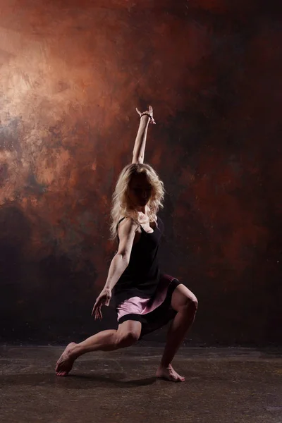 Foto av ung dansande blond kvinna med upphöjda armar i svart klänning på brun bakgrund — Stockfoto