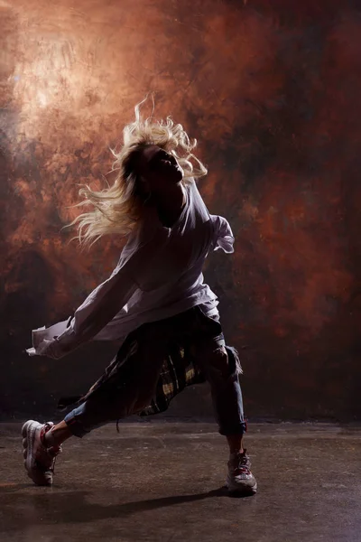 어두운 갈색 배경에 찢어진 청바지와 운동화에 슬림 금발 댄서 여자의 사진 — 스톡 사진