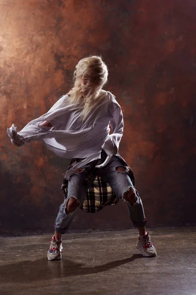 Foto de bailarina rubia de pelo largo con el pelo en desarrollo en jeans rotos y zapatillas de deporte sobre fondo marrón oscuro —  Fotos de Stock