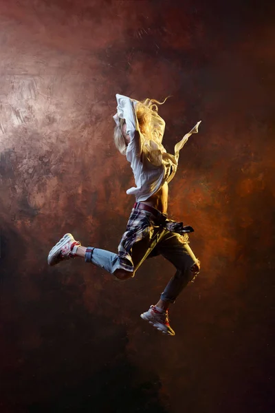 Foto de dançarina loira pulando olhando para trás em jeans rasgados e tênis em fundo marrom escuro — Fotografia de Stock