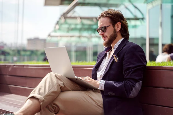 Foto auf der Seite eines jungen Geschäftsmannes mit Laptop, der an einem Sommertag auf einer Holzbank in der Innenstadt sitzt — Stockfoto