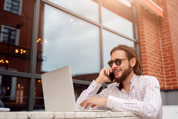 Foto di allegro uomo freelance parlando al telefono mentre seduto a tavola con computer portatile vicino edificio in mattoni — Foto Stock