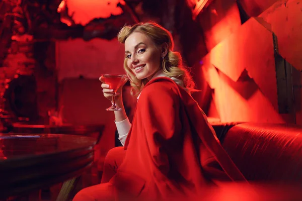 Imagen de la rubia feliz en chaqueta roja mirando a la cámara con cóctel en la mano en el club nocturno — Foto de Stock