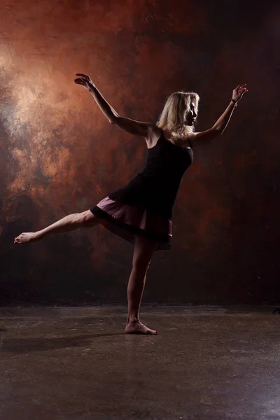 Foto de comprimento total de dança menina loira olhando para o lado em vestido preto curto no fundo marrom — Fotografia de Stock