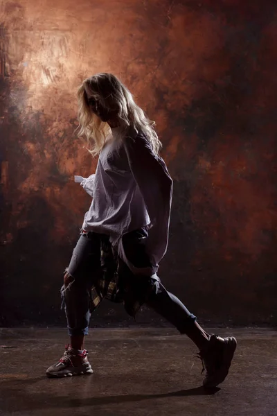 Foto lateral de dançarina loira magra em jeans rasgados e tênis em fundo marrom escuro — Fotografia de Stock