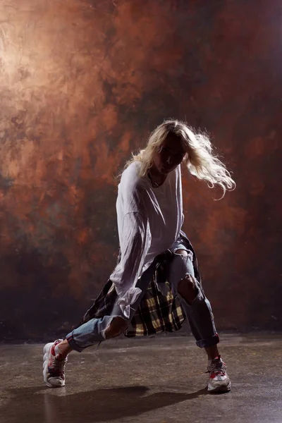 Image of long-haired blonde dancer with developing hair in torn jeans and sneakers on dark brown background — Stock Photo, Image